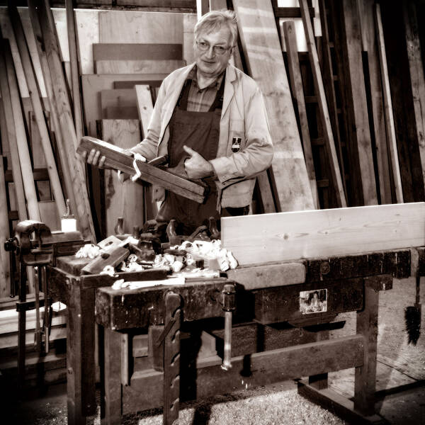Ein Tischler in seiner Werkstatt mit einem alten hölzernen Hobel hinter der Werkbank. im Hintergrund stehen Bretter. Schwarz-Weiß.