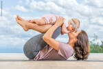 Eine Frau liegt an einem Strand auf einer Matte am Rücken und hält auf ihren angewinkleten Beinen ein Kleinkind, das sie küsst.