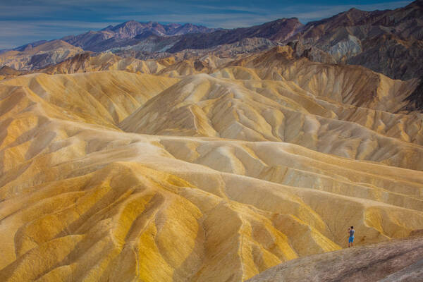 Ein Mann in blauer Short steht vor den goldgelben, sandigen Hügeln von Zabriskie Point, Death Valley.