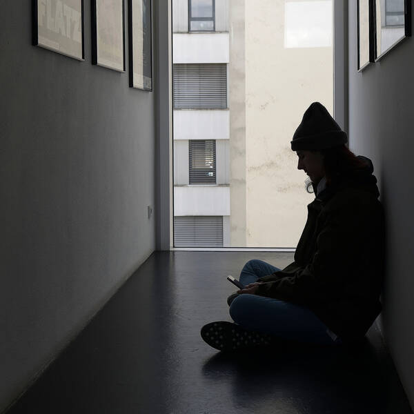 Silhouette eines Mannes mit Mütze, der im Gang eines mehrgeschoßigen Hauses sitzend auf sein Mobiltelefon blickt.