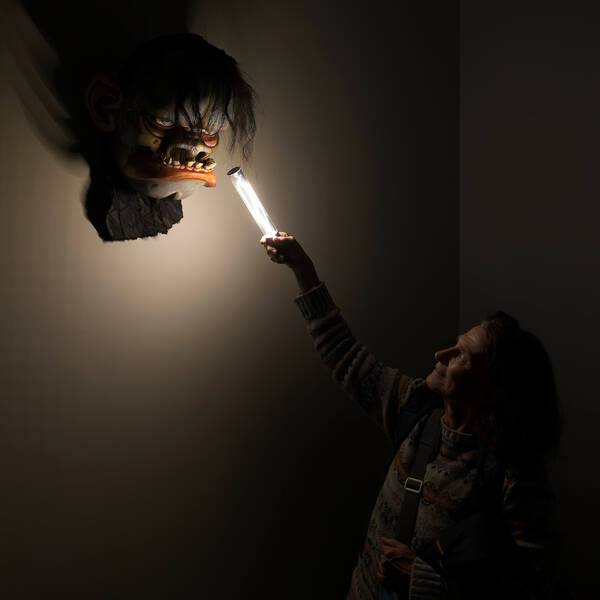 Eine Frau in einem dunklen Raum hält eine stabförmige Lampe nach oben und beleuchtet damit eine geschnitzte Maske an der Wand.