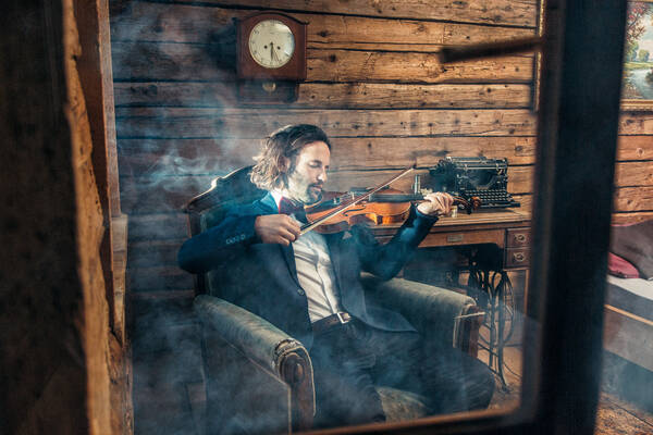 Blick durch ein Fenster in einen grob gezimmerten, altmodisch eingerichteten, verrauchten Raum. In einem Lehnsessel sitzt ein Mann mit Jackett und Fliege der auf einer Geige spielt.