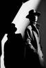 Ein Mann im Trenchcoat mit Hut im Stil des film noir mit ausgeprägten Schatten. Schwarz-Weiß.