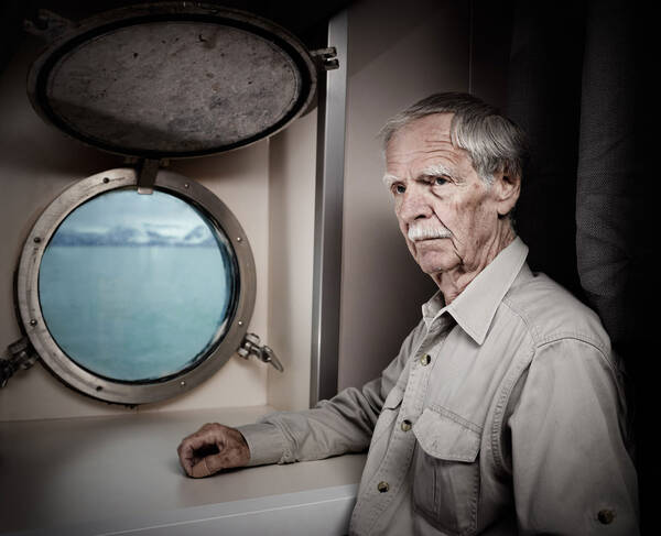 Ein älterer Herr sithe neben einem Bullauge eines Schiffes.