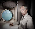 Ein älterer Herr sithe neben einem Bullauge eines Schiffes.
