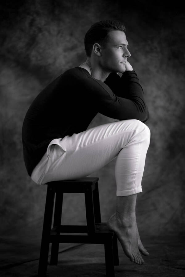 Ein junger Mann sitzt auf einem Hocker und schaut nachdenklich in die Ferne. Schwarz-Weiß.