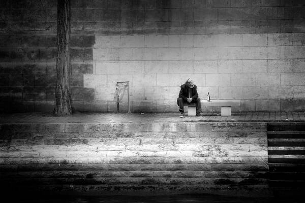 Ein alter Mann mit Kopfhörern sitzt neben einer Weinflasche auf einer Bank vor einer Steinmauer. Schwarz-Weiß.