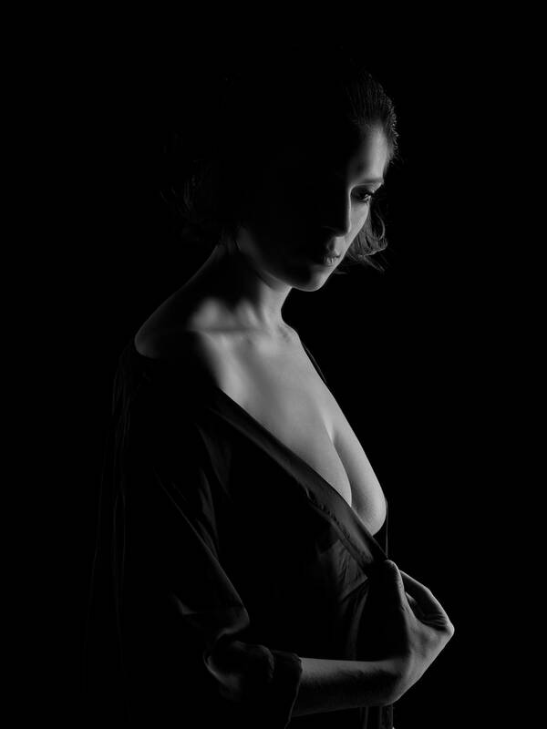 Low-Key-Portrait einer jungen Frau, die gerade ihr Gewand über die Schulter gleiten lässt. Licht kommt von rechts hinten. Schwarz-Weiß.
