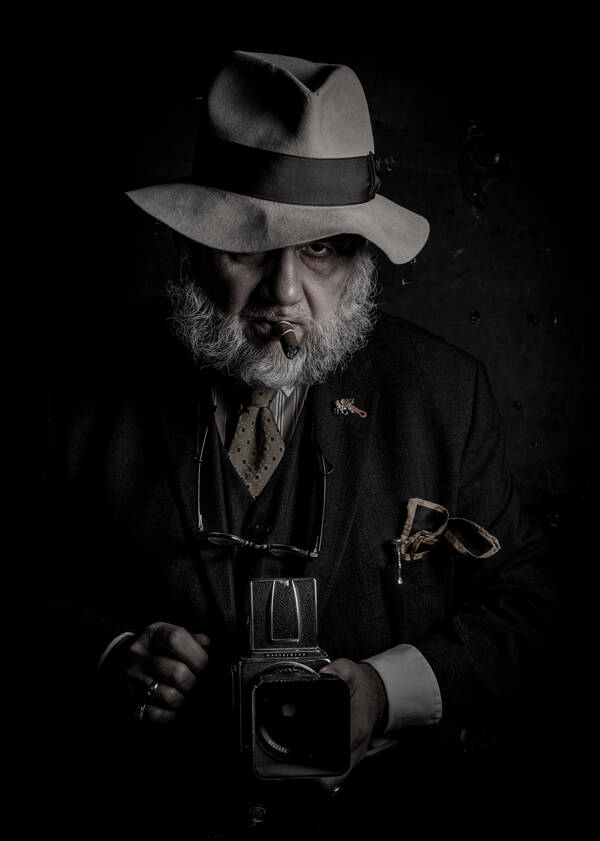 Portrait des Fotografen Robert W. Sackl-Kahr Sagostin mit Hut, Zigarre und einer antiquarischen Hasselblad-Kamera. Low Key.