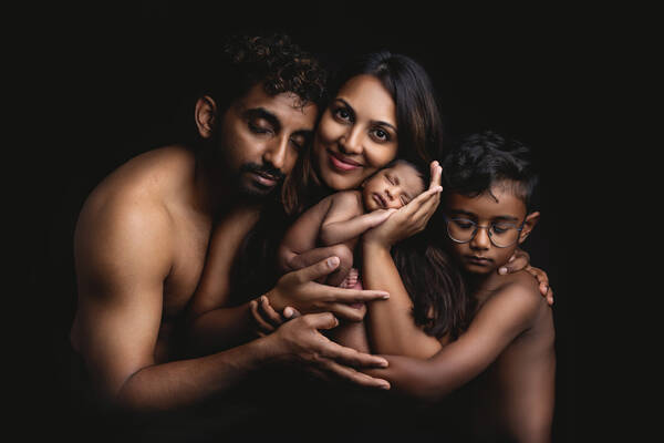 Low-Key-Aunfahme einer dunkelhäutigen Familie, die eng aneinandergeschmiegt ist. Die Mutter hat en Neugeborenes in der Hand. Der Vater und das öltere Kind umfassen mit den Händen Mutter und Baby. Außer der Mutter, die direkt in die Kamera schaut, haben alle die Augen geschlossen.