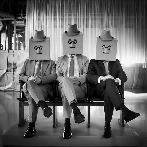 Drei Männer sitzen in Anzügen auf Kinosesseln in einem großen Saal. Über den Köpfen haben sie Kartonschachteln gestülpt, auf die Gesichter gemalt sind. auf den Kartonschachteln sitzen Hüte. Die Männer haben die Hände gefaltet in den Schoß gelegt und die Beine überkreuzt. Schwarz-Weiß.