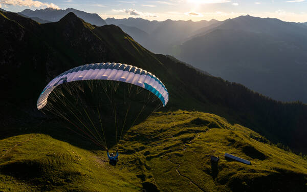 Ein Gleitschirmflieger über einem Almboden in den Bergen im Abendlicht. unter ihm sieht man eine Alm.