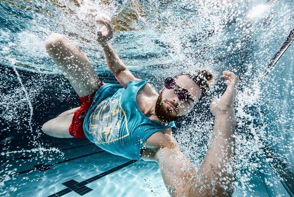 Ein bärtiger Mann in Shorts und Shirt taucht in ein Schwimmbecken ein. Unterwasseraufnahme.