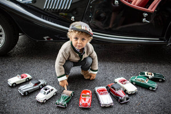 Ein kleiner, blondgelockter Bub mit Schirmmütze hockerlt vor einem eleganten Oldtimer und hat vor sich im Halbkreis aufgereihte Oldtimer-Modell-Autos aufgereiht.
