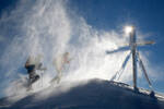 Zwei Personen in Bergausrüstung mit Stöcken und Schneeschuhen nähern sich einem Gipfelkreuz durch den vom Wind aufgewirbelten Schnee im Gegenlicht.