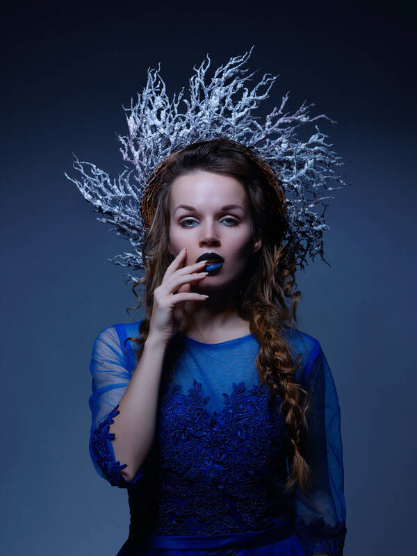 Portrait einer jungen Frau in blauem, onamentalen Kleid und mit blau geschminkten Lippen. die langen brünetten Haare bilden starke, unregelmäßige Zöpfe. am Kopf hat sie einen Kranz, der an weiße Korallen erinnert.