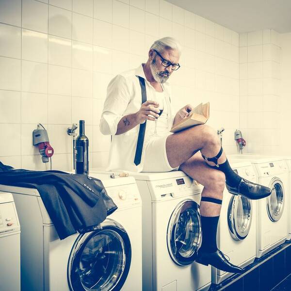 In einem weiß gefliesten Waschsalon sitzt ein eleganter, reiferer Herr mit Bart und Brille in Unterhose auf einer der Waschmaschinen und liest bei einem Glas rotwein ein Buch.