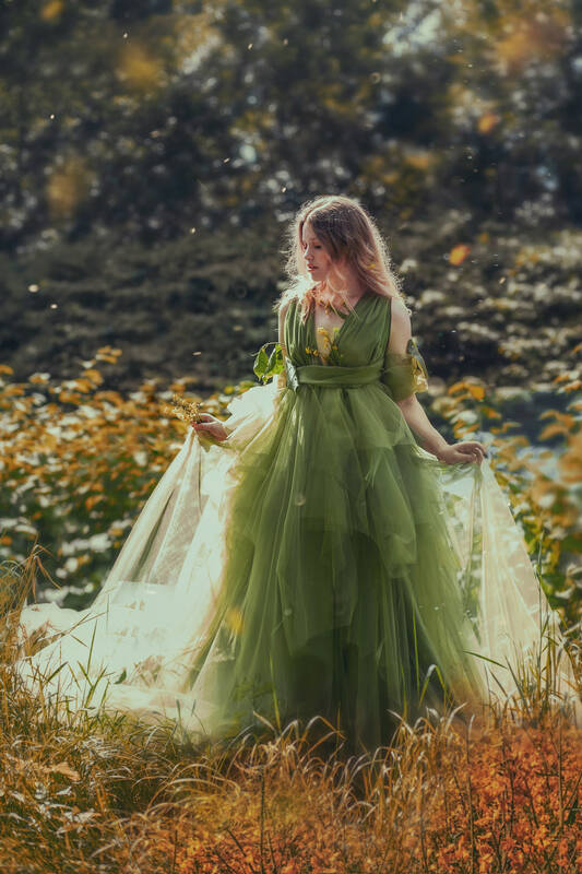 Eine junge, langhaarige Frau in einem vielschichtigen sonnendurchfluteten grünen Kleid im Grünen. In der Hand und im Ausschnitt hat sie gelbe Blumen.
