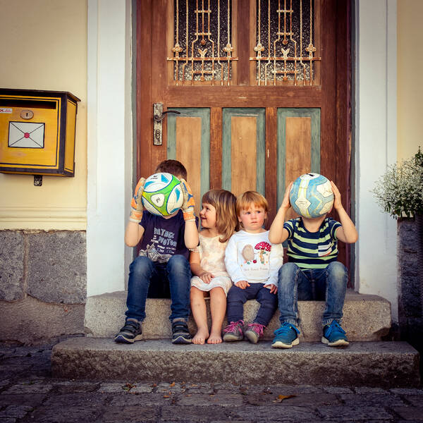 Vier Kinder sitzen auf der Treppe vor einer historischen Türe. Zwei Kinder halten einen Ball vor ihr Gesicht.