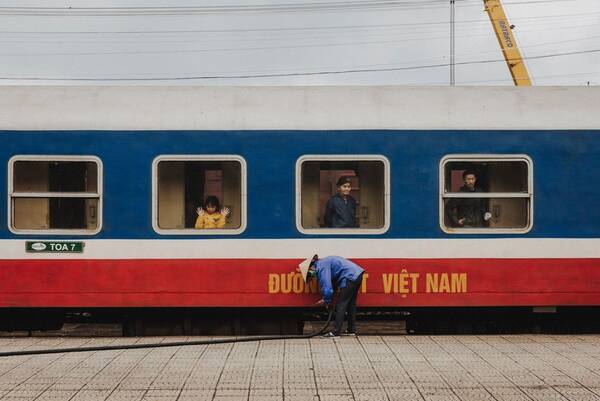 In einem Waggon der Đường sắt Việt Nam (Nationale Eisenbahn Viet Nam) stehen ein Mädchen, eine alte Frau und ein Mann je an einem Fenster während ein Arbeiter einen Wasserschlauch ansteckt.