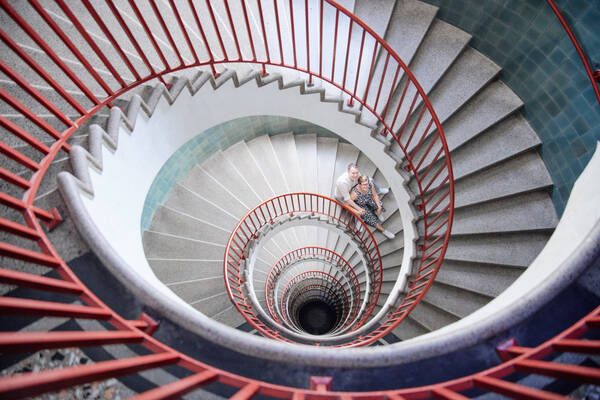 Blick von oben in das markante Stiegenhaus des Nebotičnik-Turmes mit dem auffälligen roten Stiegengeländer und den hellblau gefliesten runden Wänden. etwa eineinhalb Stockwerke tiefer sitzt ein Paar auf der Stiege und schaut nach oben in die Kamera