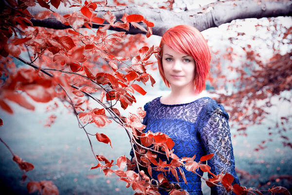 Eine junge Frau mit kurzen roten Haaren steht mit dunkelblauen Kleid in einem Park hinter einem Rotbuchenzweig.
