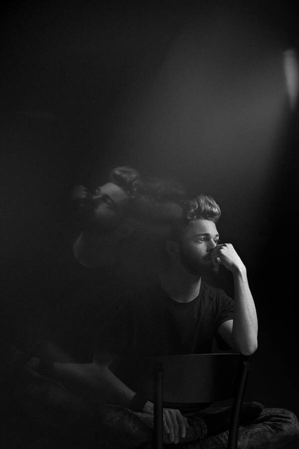 Ein bärtiger junger Mann mit nachdenklichem Ausdruck sitzt im Schneidersitz rittlings auf einem Sessel, ein Ellenbogen auf die Sessellehne gestützt. Das Bild ist in einer Diagonale gespiegelt. Schwarz-Weiß.