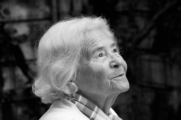 Portrait einer alten, weißhaarigen Frau, die nach rechts oben aus dem Bild blickt. Schwarz-Weiß.