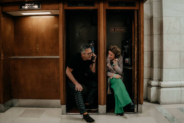 Ein Mann und eine Frau sitzen in historischen hölzernen Telefonzellen und telefonieren und schauen sich dabei an.