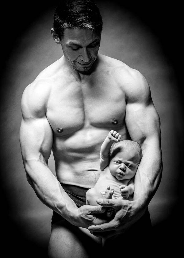 Ein sehr muskulöser Mann formt mit seinen Armen einen hängenden Bogen in dem ein Neugeborenes liegt. Starke Vignettierung. Schwarz-Weiß.