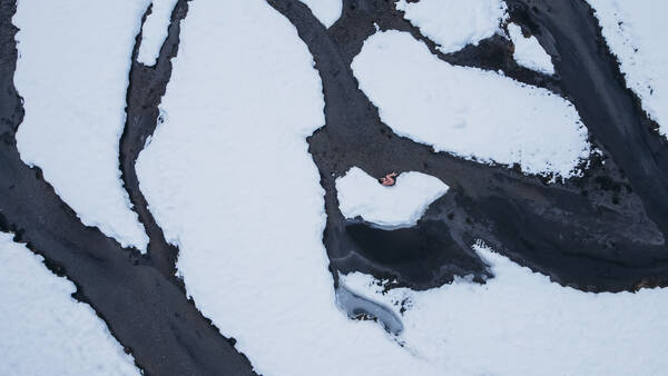 Eine nackte Frau liegt in Embryo-Stellung in einem Bachbett in verschneiter Landschaft. Dronenfoto.