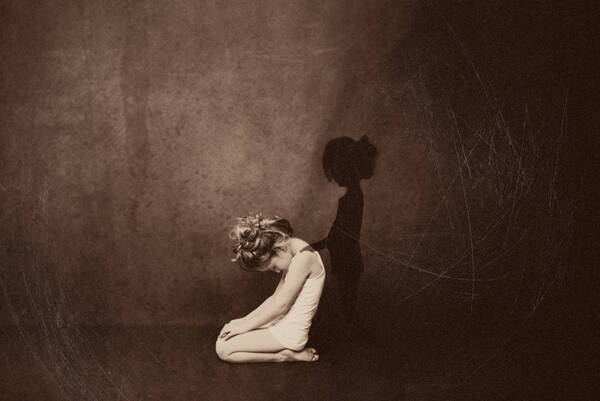 Ein kleines Mädchen mit blondem Wuschelkopf im Unterhemd kniet mit gesenktem Kopf. Der Schatten eines Mädchens steht neben ihr und legt ihr die Hand auf die Schulter.