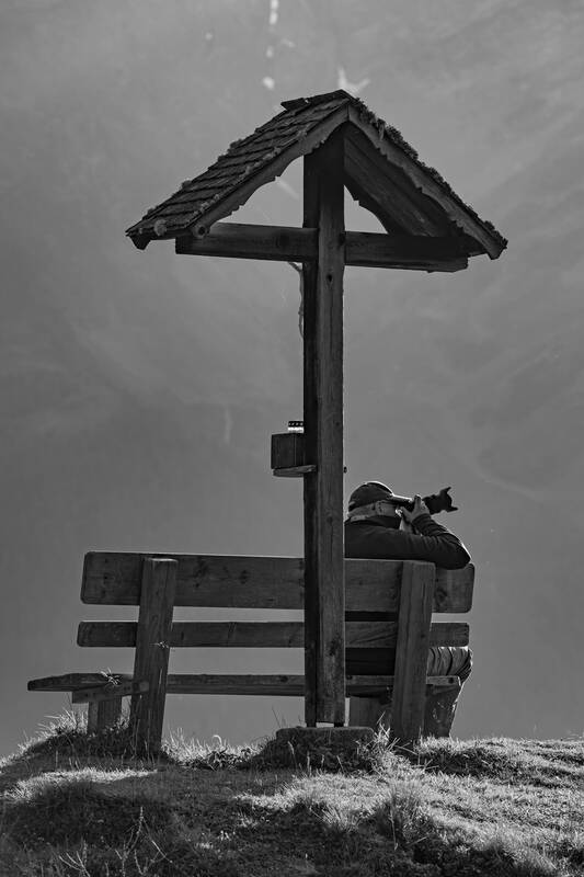 Ein älterer Mann sitzt bei einem Wegkreuz im Gebirge auf einer Bank und Fotografiert. Schwaraz-Weiß.