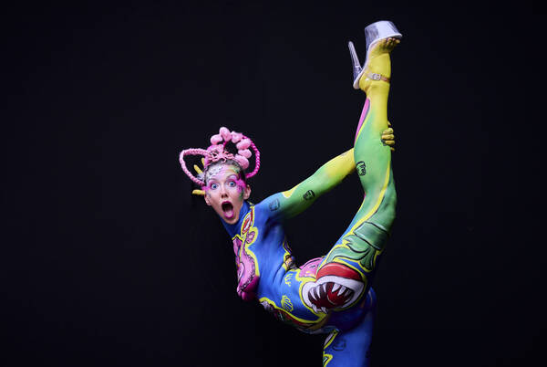 Eine in lebhaften Faben bemalte Frau in einer akrobatischen Pose vor schwarzem Hintergrund. 