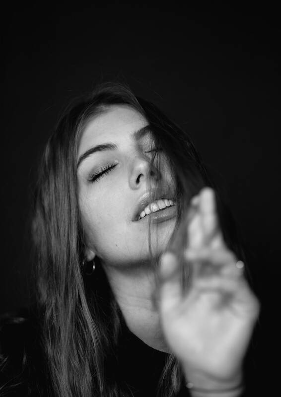 Portrait einer jungen Frau, das Gesicht wird zum Teil von den langen Haaren verdeckt. Die Augen sind geschlossen, der Mund leicht geöffnet. im Vordergrund macht die Hand eine Geste. Schwarz-Weiß