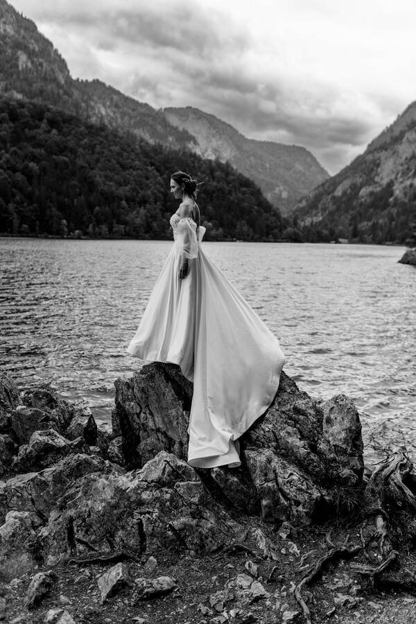 Eine Braut steht im Brautkleid auf Steinen am Ufer eines Sees in den Bergen. Schwarz-Weiß.