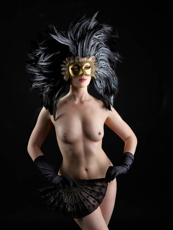 Eine junge Frau nur mit schwarzen Handschuhen und einer gold-schwarzen Federmaske bekleidet, mit den Händen hält sie einen schwarzen Fächer vor den Schambereich.
