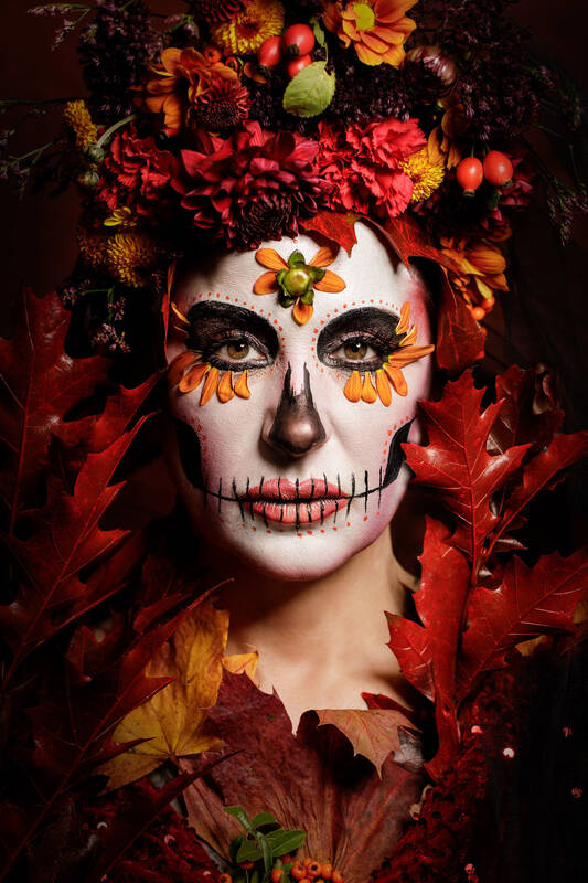 Eine Frau, deren Gesicht zum totenkopf geschminkt ist und die ganz von herbstlichen Accessoirs wie bunten Blättern, Frühchten und Blüten umgeben ist.