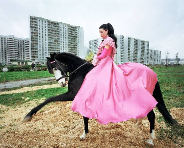 Eine junge Frau in rosanem Kleid mit Schleppe reitet vor einer russischen Plattenbau-Siedlung auf einem schwarzen Pferd.