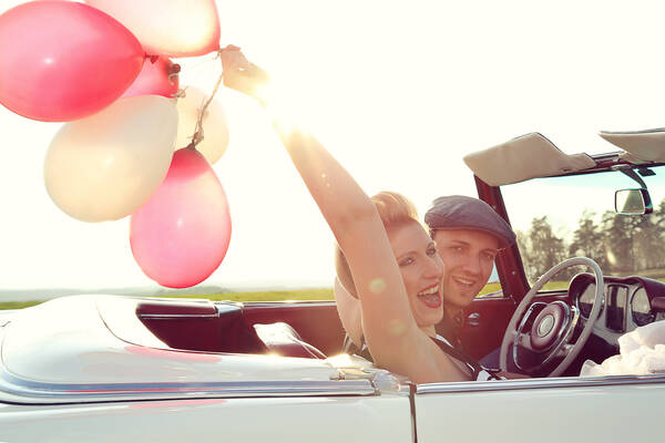 Ein Paar mit fährt im offenen Oldtimer Cabrio in der Abendsonne. Die Frau hat bunte Luftballons in der Hand.