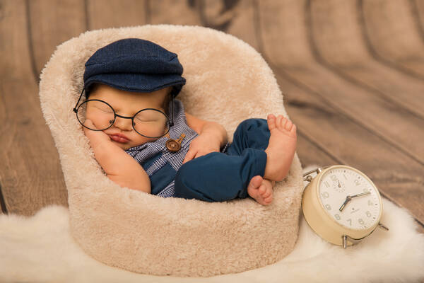 Ein schlafendes Baby mit Brille und Käppi in einem kuscheligen Sessel neben einem Wecker.