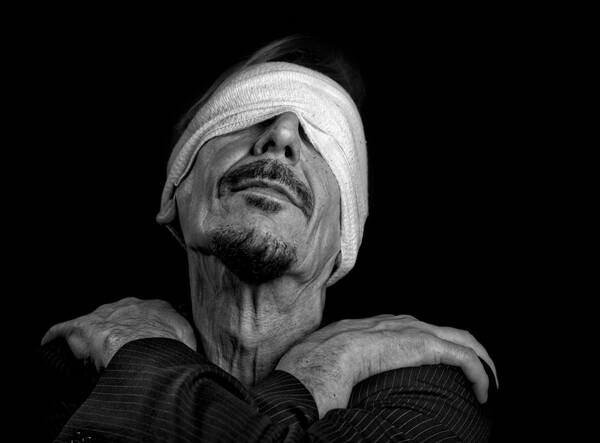 Ein Mann mit Verbundenen Augen, die Hände über Kreuz auf den eigenen Schultern. Schwarz-Weiß