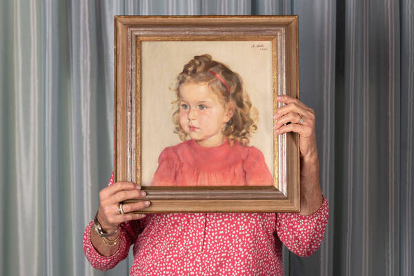 Eine ältere Frau in einer rot-weiß gemusterten Bluse hält ein Gemälte eines kleinen, blondgelockten Mädchens in rotem Kleid direkt vor ihr Gesicht.