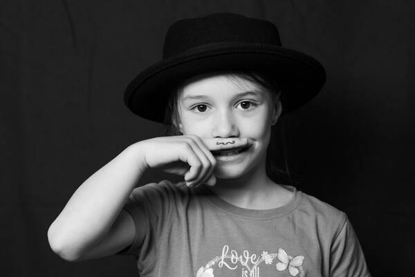 Portrait eines Kindes mit schwarzem Hut vor schwarzem Hintergrund. Das Kind hält sich den Zeigefinger vor die Oberlippe, auf den ein kleiner Schnurrbart gezeichnet ist. Schwarz-Weiß.