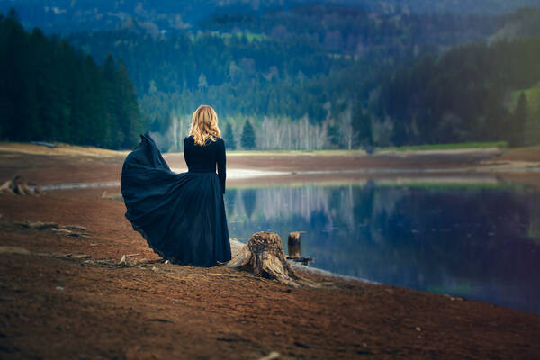 Eine Frau in dunklem Kleid steht am Ufer eines Sees. 