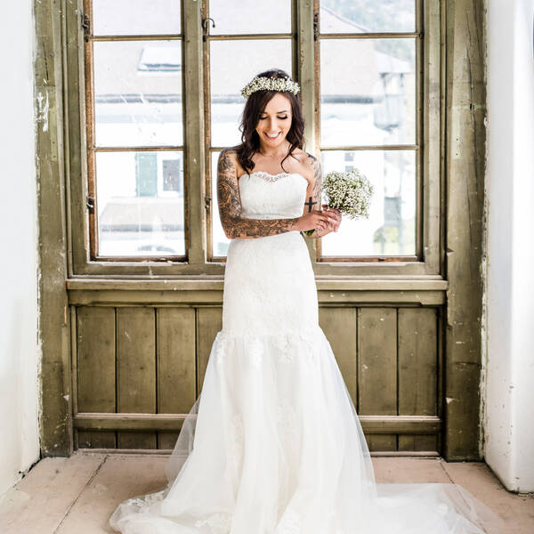 Eine Braut mir weißem Blumenkranz, zartem, kleinblütigem weißem Brautstrauß  und tätowierten Armen steht in einem alten, in die Jahre gekommenen Raum vor einer olivgrünen Holzwand mit Fenster.