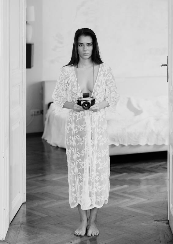 Eine junge Frau steht barfuß in weißem teiltransparentem Kleid mit einem Photoaparat in einer Wohnung. Schwarz-Weiß.