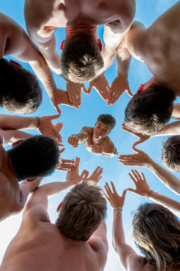 Blick von unten in eine Gruppe junger Menschen, alle mit nacktem Oberkörper, die im Kreis stehen und ihre Hände nach oben strecken. Im Zentrum des Kreises sieht man einen jungen Mann der weiter oben in Superman-Pose auf die ausgestreckten Hände 