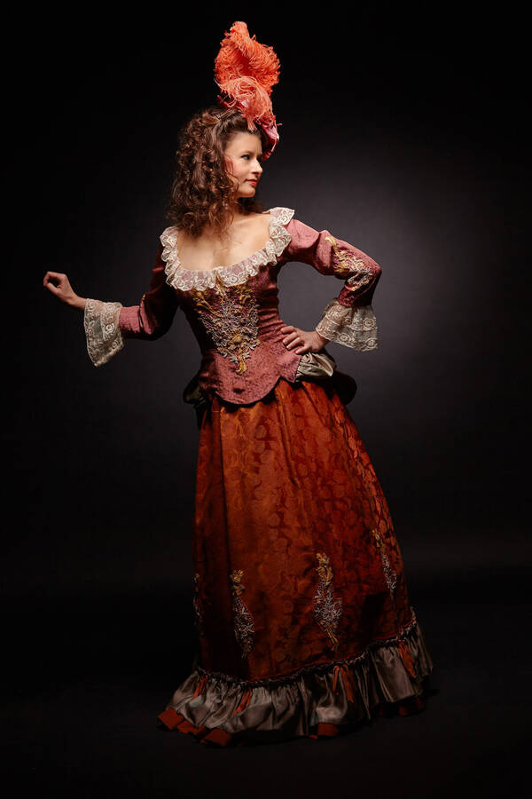 Ganzkörper-Portrait einer Frau in verspieltem, historisch anmutendem Kleid in Rot- und Brauntönen. Sie trägt einen ausgefallenen orangen Fascinator.