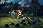 Ein Mann und eine Frau sitzen rotweintrinkend vor einer Gartenlaube im Dämmerlicht. In der Wiese liegen viele schwarze Hüte.
